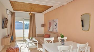 A modern living room with a sofa, dining table, and chairs. There is a large window with curtains, a ceiling with a woven design, and a wall-mounted mirror.