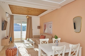 A modern living room with a sofa, dining table, and chairs. There is a large window with curtains, a ceiling with a woven design, and a wall-mounted mirror.