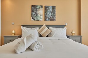A neatly made bed with white linens and a rolled towel, two nightstands with decorative lamps, and two wall-mounted artworks depicting foliage.