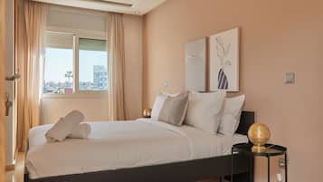 A neatly made bed with white linens and a rolled towel, a black bedside table with a decorative golden sphere, and two abstract wall art pieces.