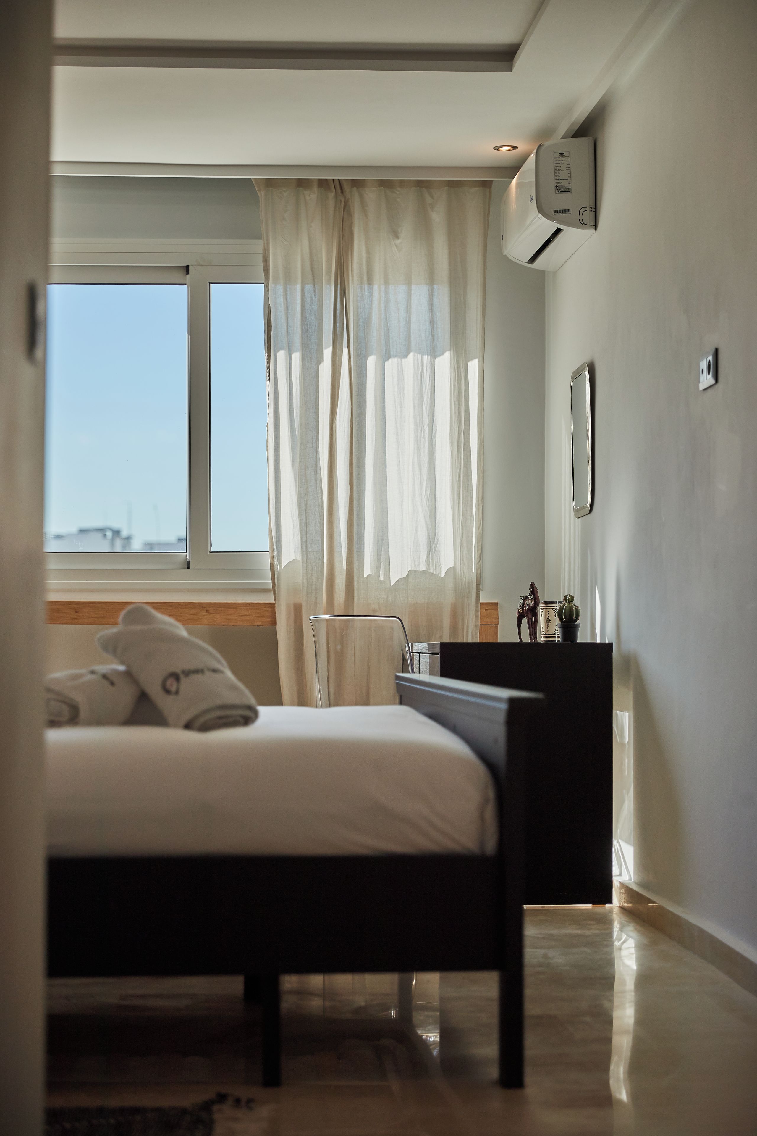 A bedroom with a bed, a window with curtains, a bedside table, and an air conditioning unit.