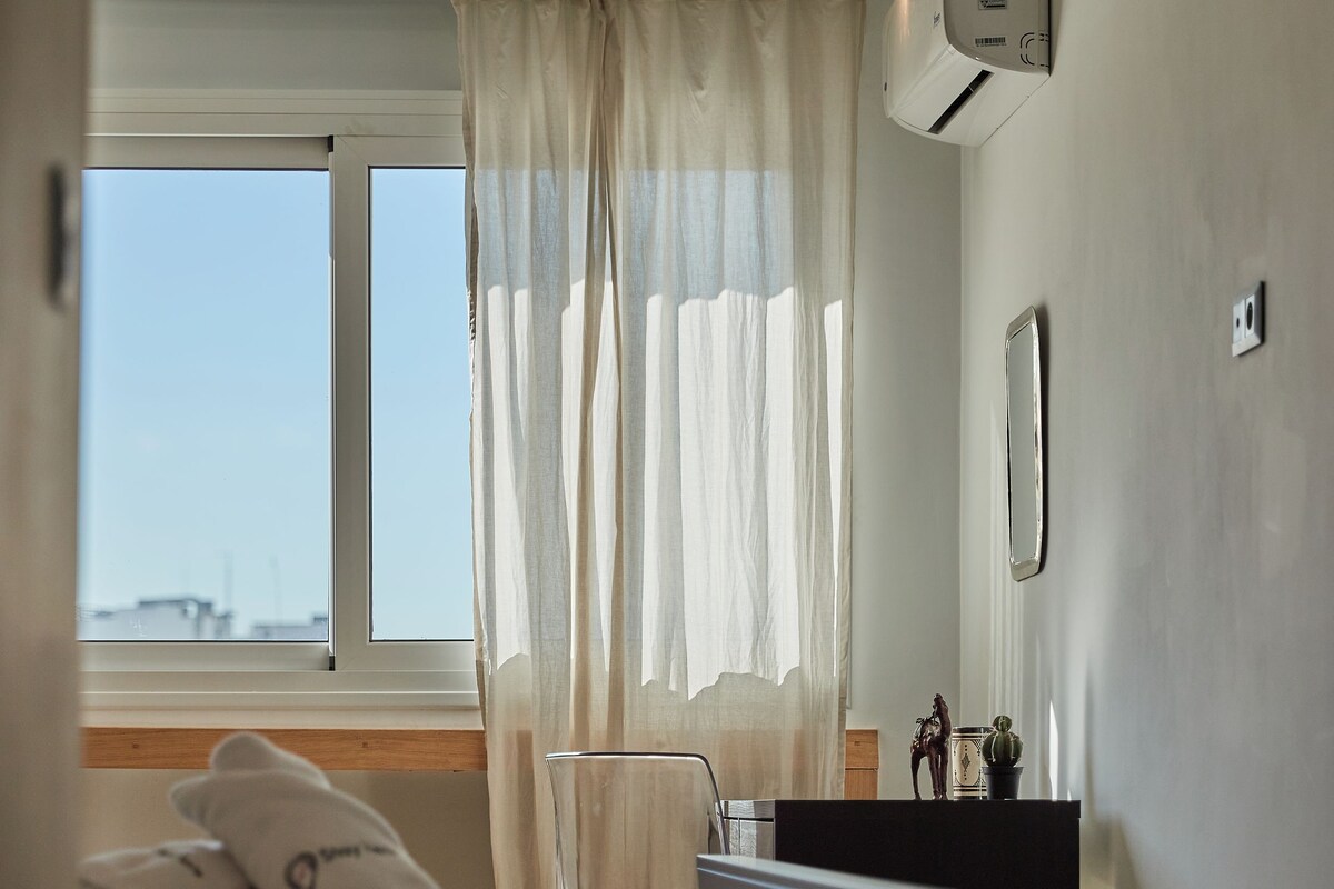 A bedroom with a bed, a window with curtains, a bedside table, and an air conditioning unit.