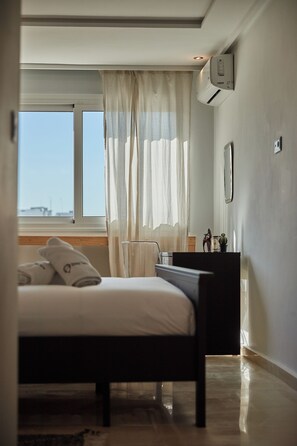 A bedroom with a bed, a window with curtains, a bedside table, and an air conditioning unit.
