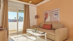 A modern living room with a sofa, ottoman, and coffee table. There is a large window with curtains, a ceiling with a geometric pattern, and artwork on the wall. - Stayhere Rabat - Hassan Heritage (Rabat)