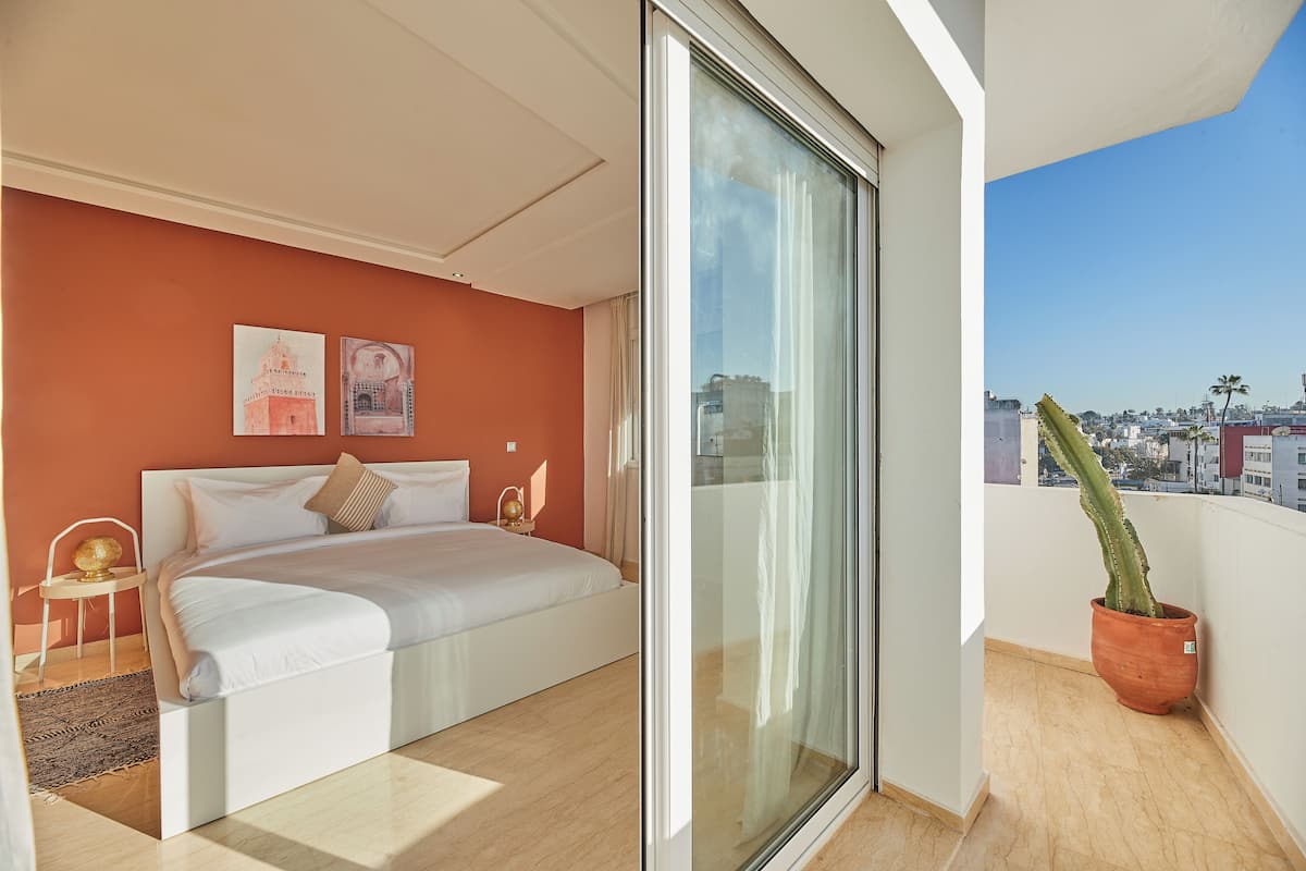 A modern bedroom with a large bed, a glass door leading to a balcony, and a city view.