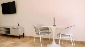 Een moderne kamer met een ronde witte tafel en twee grijze stoelen, een flatscreen televisie aan de muur bevestigd en een klein witte bijzettafeltje met een lamp.