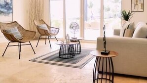 Een ruime woonkamer met een groot raam, een beige bank, twee rieten fauteuils, een salontafel en een vloerlamp.