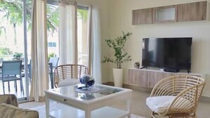 Una sala de estar con una mesa de centro blanca, sillas de mimbre, un televisor y vistas a palmeras afuera.