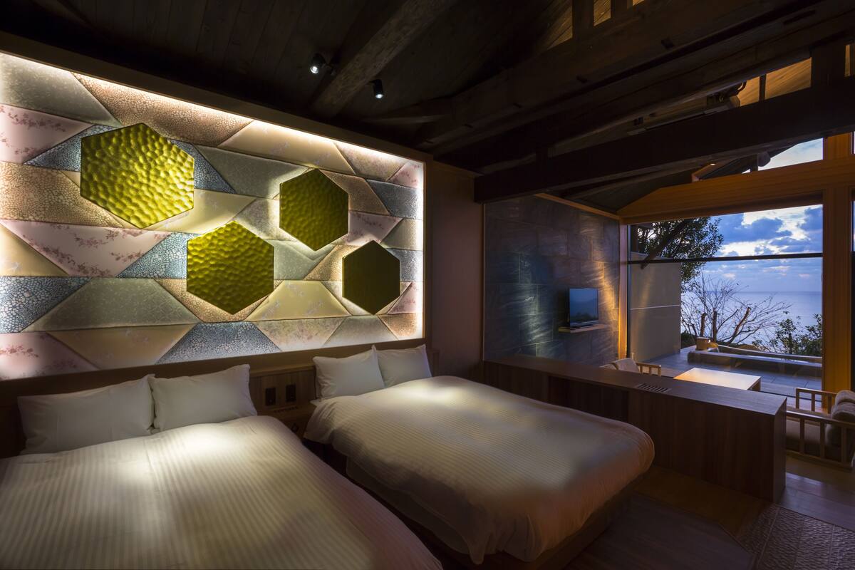 A modern hotel room with two beds, a large window with a view, and a unique hexagonal wall design.