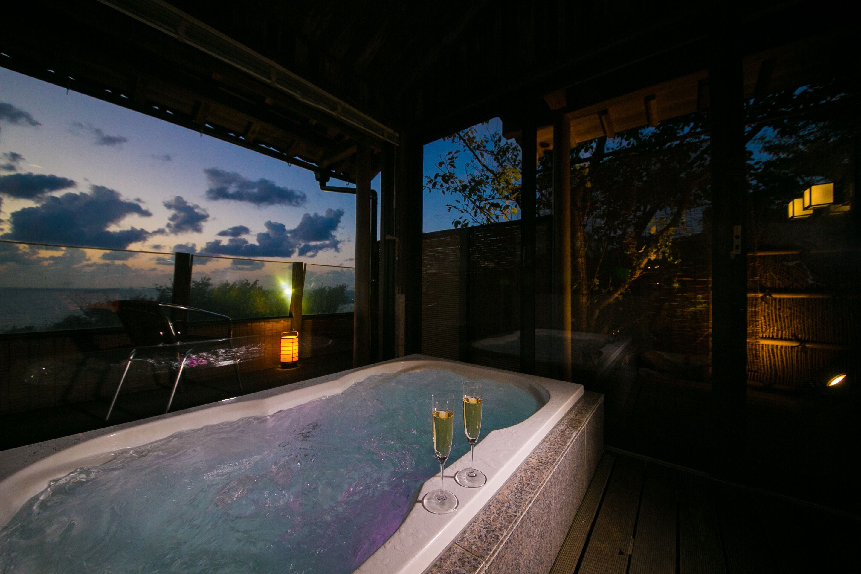 A hot tub with two glasses of champagne, a wooden deck, and a view of the sky and trees.