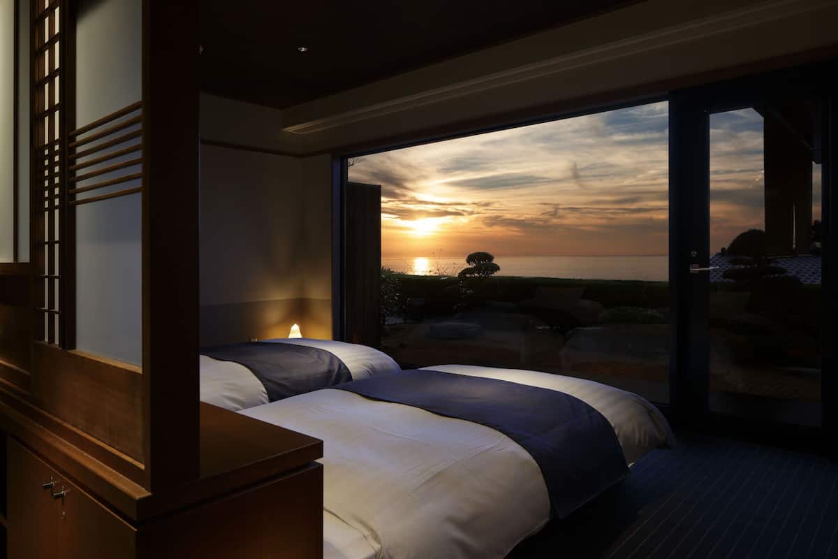 A bedroom with a large bed, a wooden dresser, and a view of the sunset through a glass door.