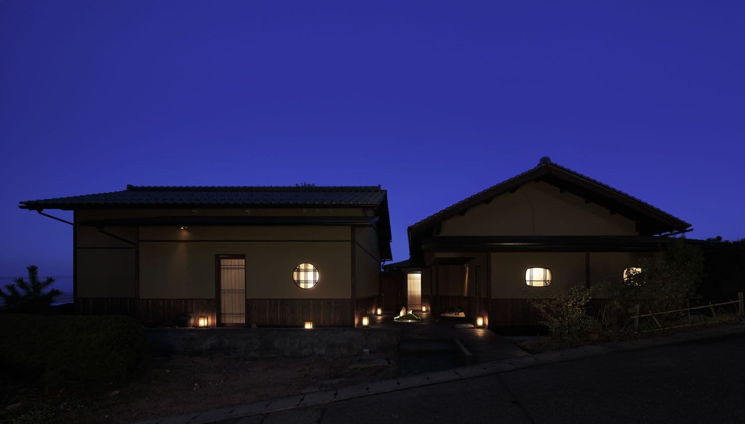Deux maisons traditionnelles de style japonais, reliées à l’entrée par un sentier de pierres et illuminées par de petites lanternes.