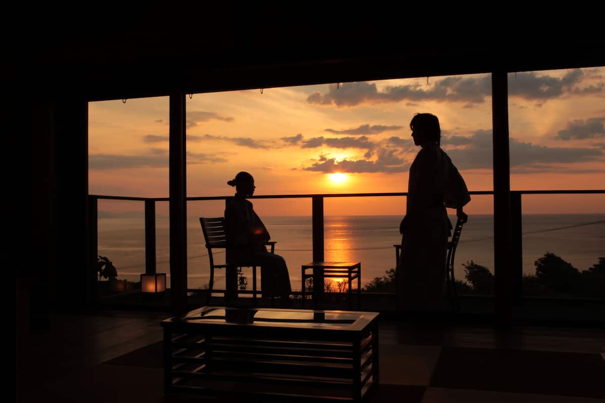 A room with large windows overlooking a sunset. There are two chairs and a small table in the foreground.