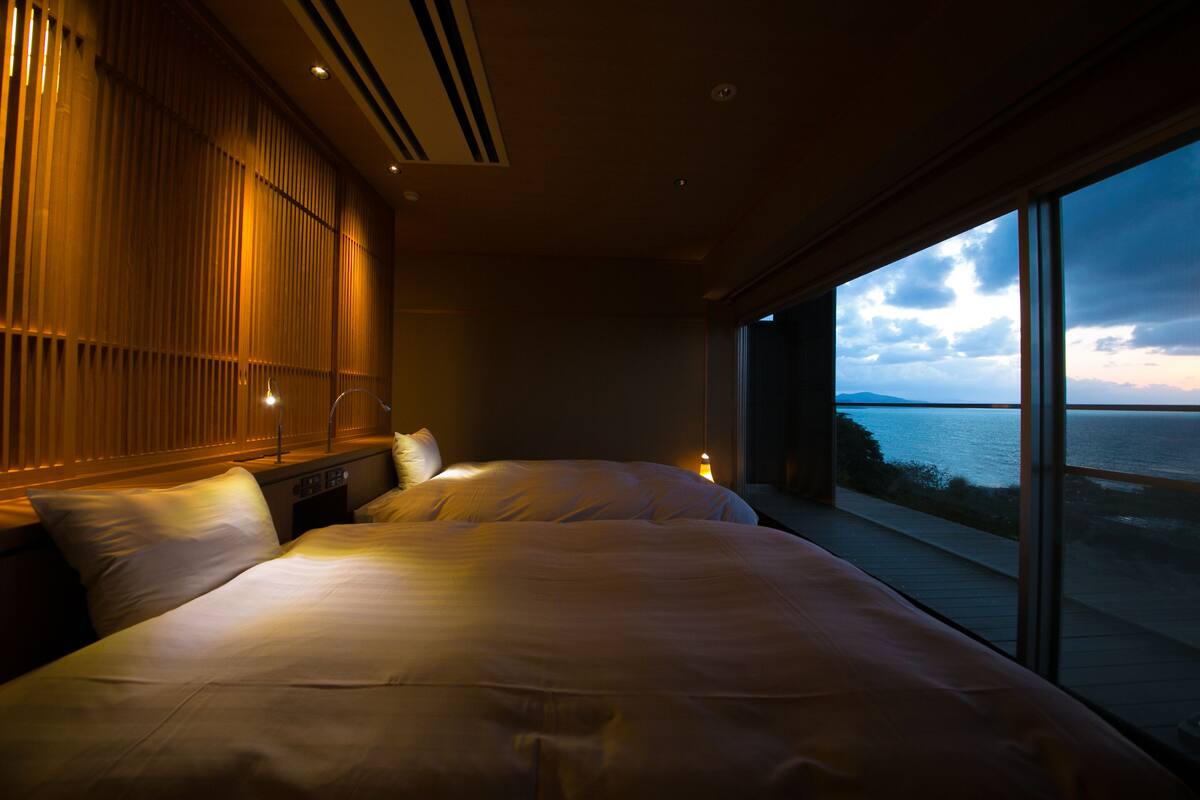 A modern bedroom with a large bed, a nightstand, and a view of the sea through large windows.
