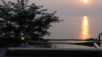 A wooden deck with a hot tub, a tree, and a view of the sunset over the water.