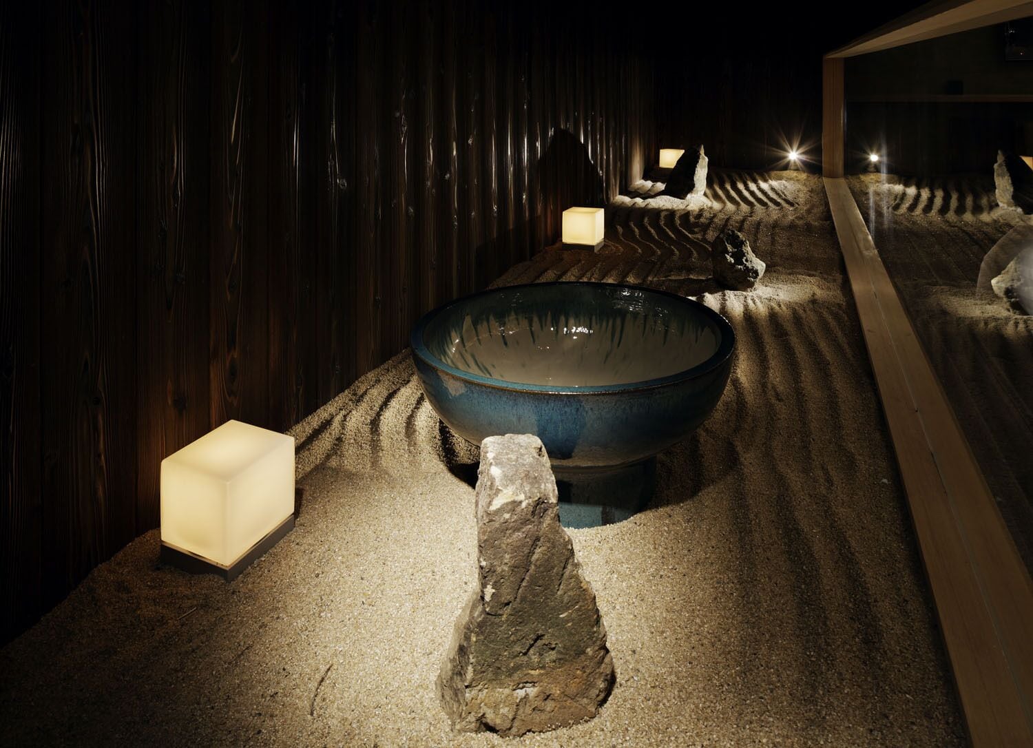 Una stanza poco illuminata con una grande ciotola blu al centro, circondata da sabbia e rocce.