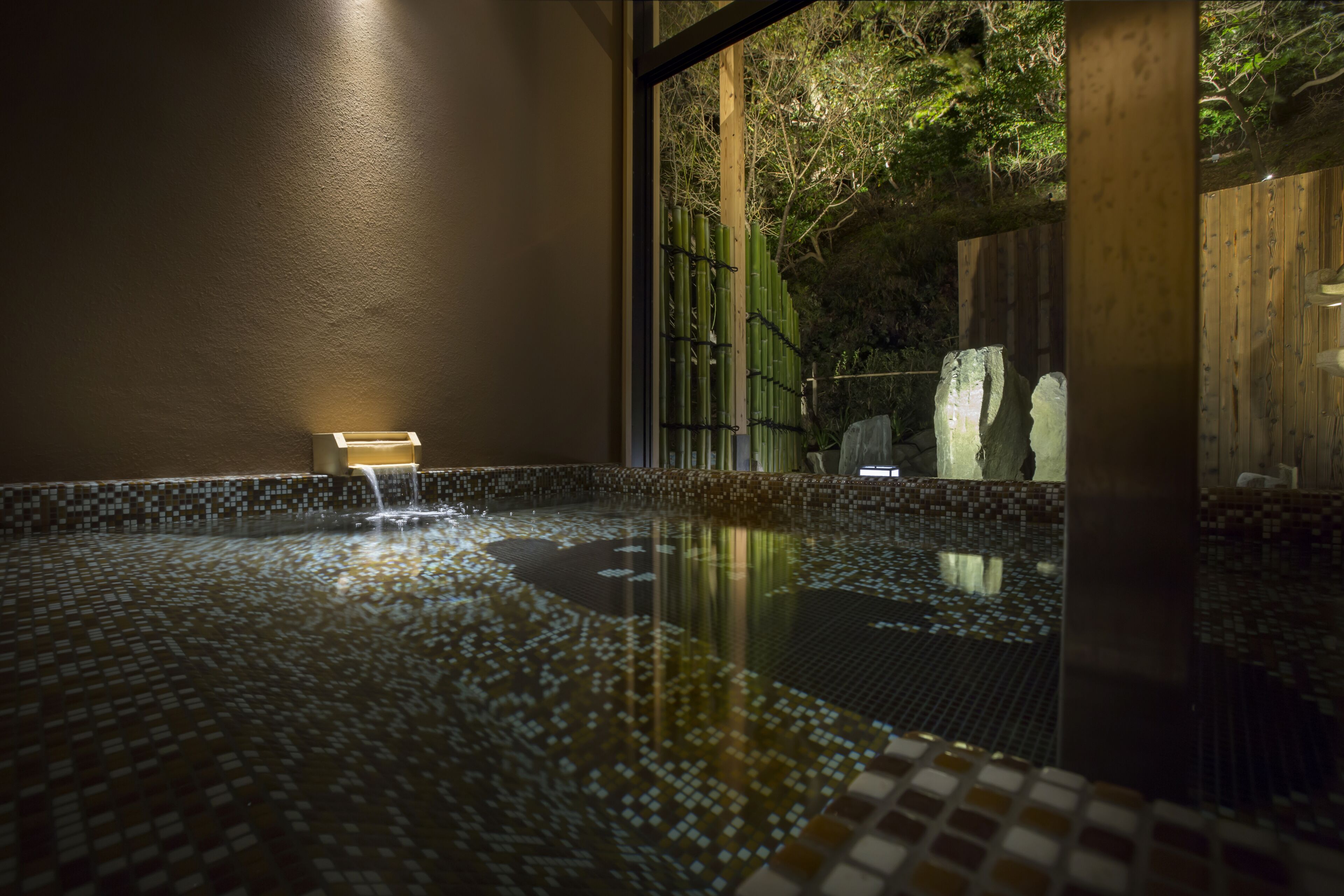 A tranquil indoor pool with a tiled floor, a bench, and a view of a wooden fence and trees outside.