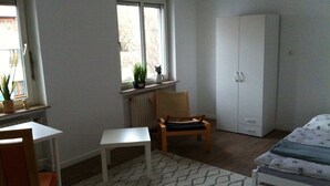 Ein Schlafzimmer mit einem Einzelbett, einem Sessel, einem kleinen Tisch, einem Kleiderschrank und einem Fenster mit einer Pflanze.