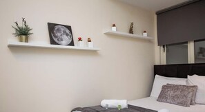 Une chambre à coucher comprenant un lit, une table de chevet, une étagère murale décorative et une fenêtre avec des stores.