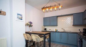 Une cuisine moderne avec des meubles bleus, une table à manger en bois et des chaises en osier, un luminaire suspendu et une fenêtre agrémentée d’un paravent décoratif.