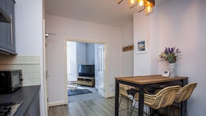 Une cuisine moderne équipée d’un four à micro-ondes, un coin repas avec une table en bois et des chaises en osier, et un couloir menant à un salon avec un téléviseur.