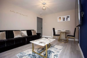 Una sala de estar con un sofá negro, una mesa baja de mármol, un comedor con una mesa y sillas blancas, y una lámpara en el techo.