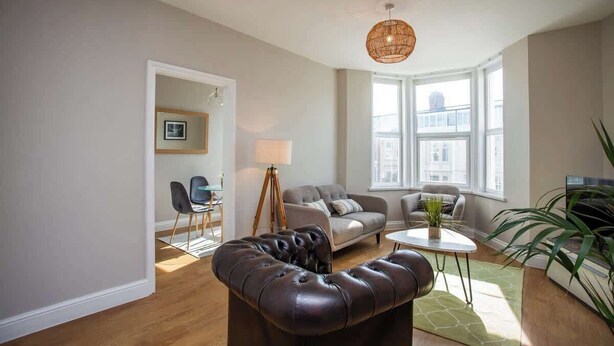 A modern living room with a leather sofa, a small table, and a lamp. There are two windows providing natural light. - Whitey Bay Coastal Bliss (Whitley Bay)