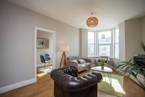 A modern living room with a leather sofa, a small table, and a lamp. There are two windows providing natural light.