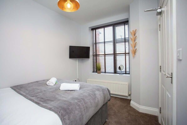A bedroom with a bed, a television, a window with blinds, and a radiator.