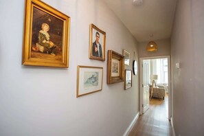 A hallway with framed artwork on the walls, a lamp, and a glimpse of a room with a bed.