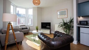 A modern living room with a sofa, a leather ottoman, a TV, a lamp, and a plant. - Whitey Bay Coastal Bliss (Whitley Bay)