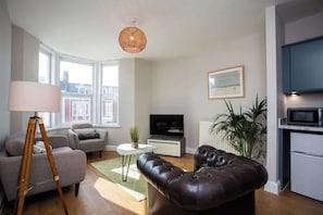 A modern living room with a sofa, a leather ottoman, a TV, a lamp, and a plant. - Whitey Bay Coastal Bliss (Whitley Bay)