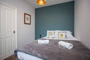 A neatly made bed with rolled towels, a framed picture on the wall, and a door to the left.