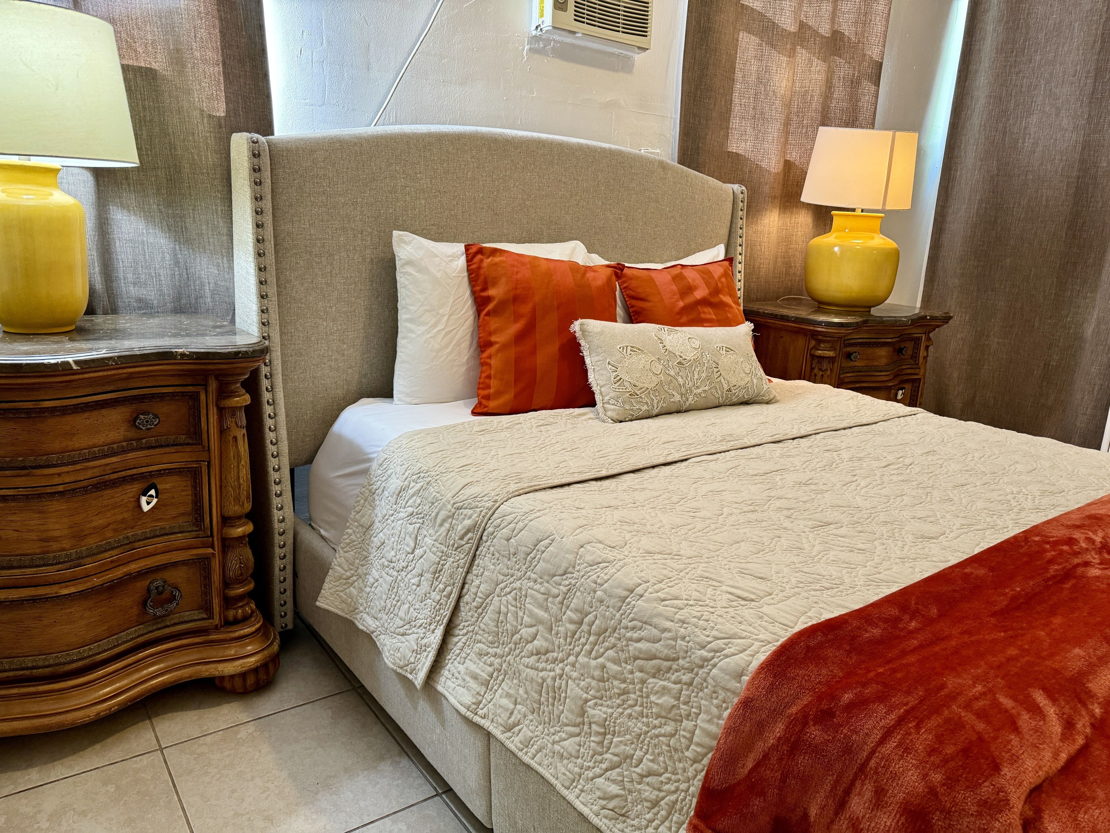 Un lit bien fait, recouvert d’une couverture matelassée, avec deux oreillers et un plaid rouge. Il y a deux tables de chevet avec des lampes, une de chaque côté du lit.