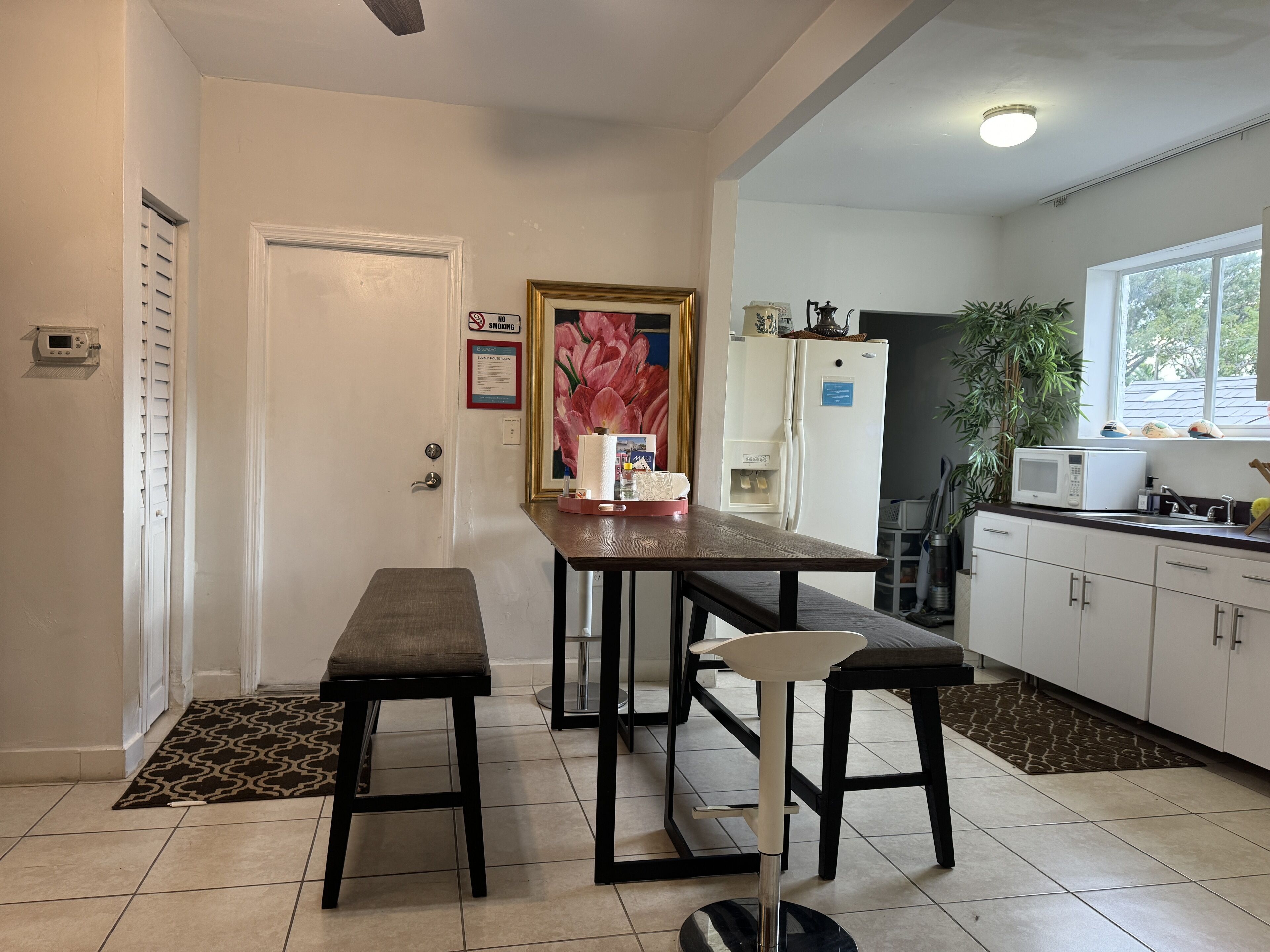 A kitchen with a dining table, two stools, a refrigerator, a microwave, and a window with a view of trees.