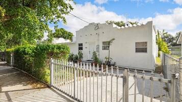 Stucco home with a flat roof and a small porch, surrounded by a picket fence and potted plants.