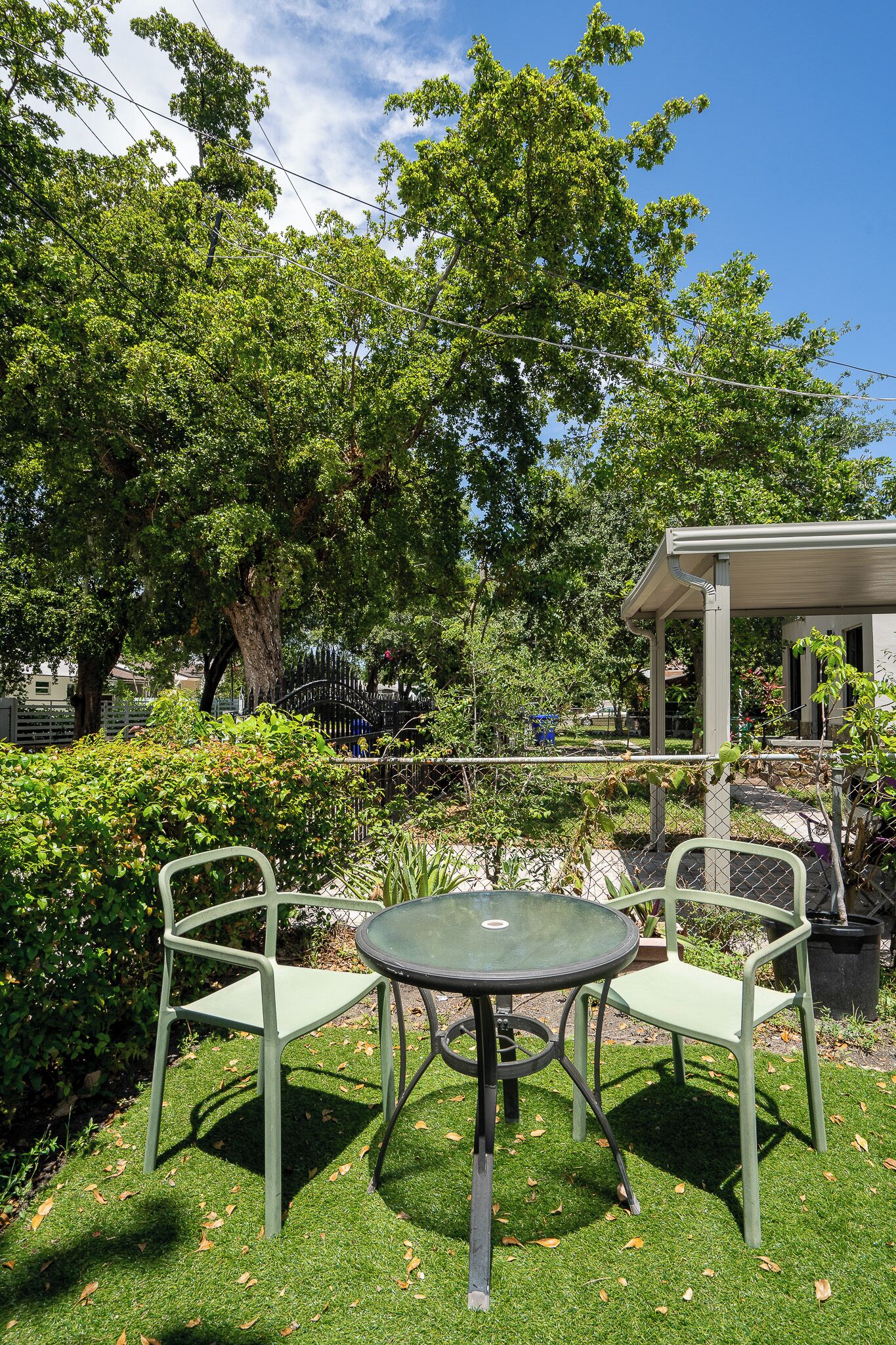Eine Terrasse mit zwei grünen Metallstühlen und einem runden Tisch, umgeben von üppiger Bepflanzung und einem großen Baum.