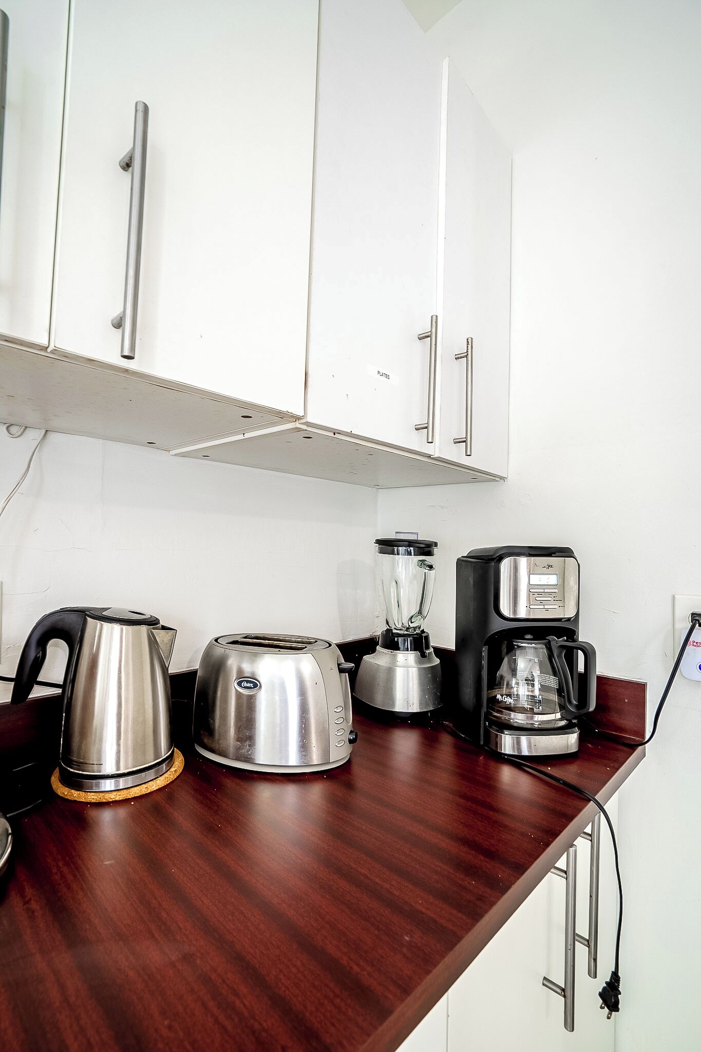 Une cuisine avec des meubles blancs, une bouilloire en acier inoxydable, un grille-pain, un mixeur et une machine à café, le tout posé sur un plan de travail en bois.