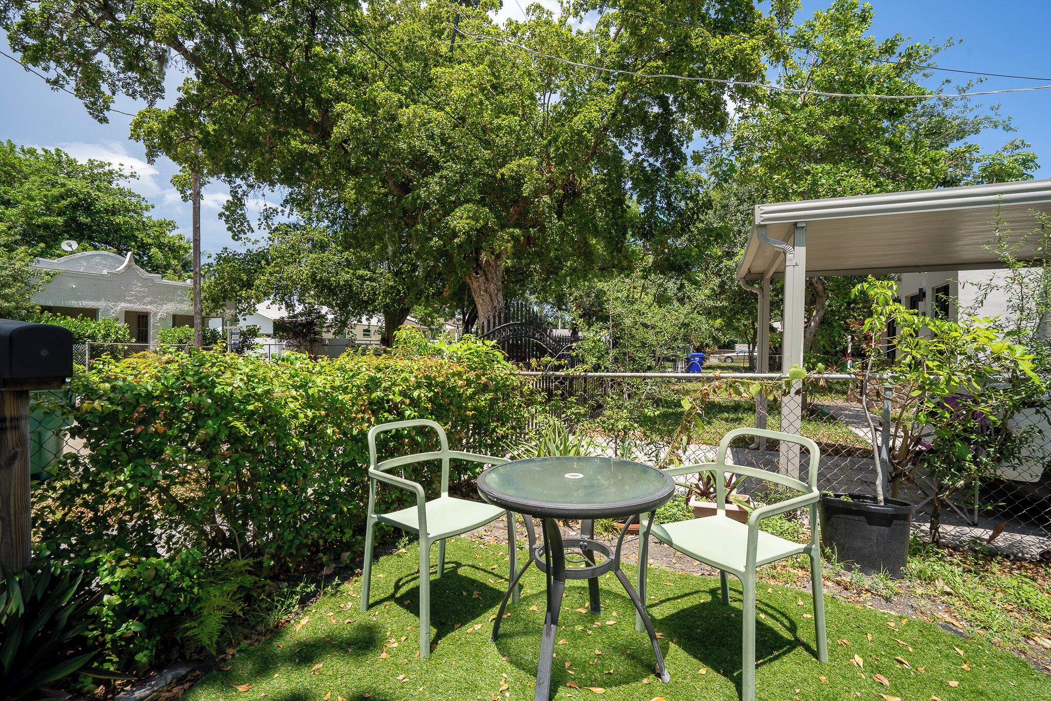 Un patio con due sedie in metallo e un tavolo, immerso nel verde e ombreggiato da un albero.