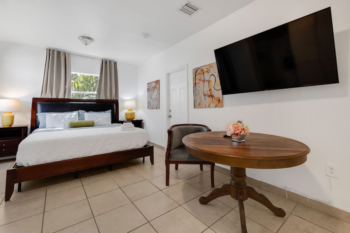 Un dormitorio con cama, lámparas de noche, una mesa redonda de madera con flores, un televisor montado en la pared y obras de arte.