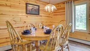 Una habitación con paneles de madera, una mesa redonda de madera, seis sillas, una lámpara colgante, un cuadro en la pared y una ventana con cortinas.