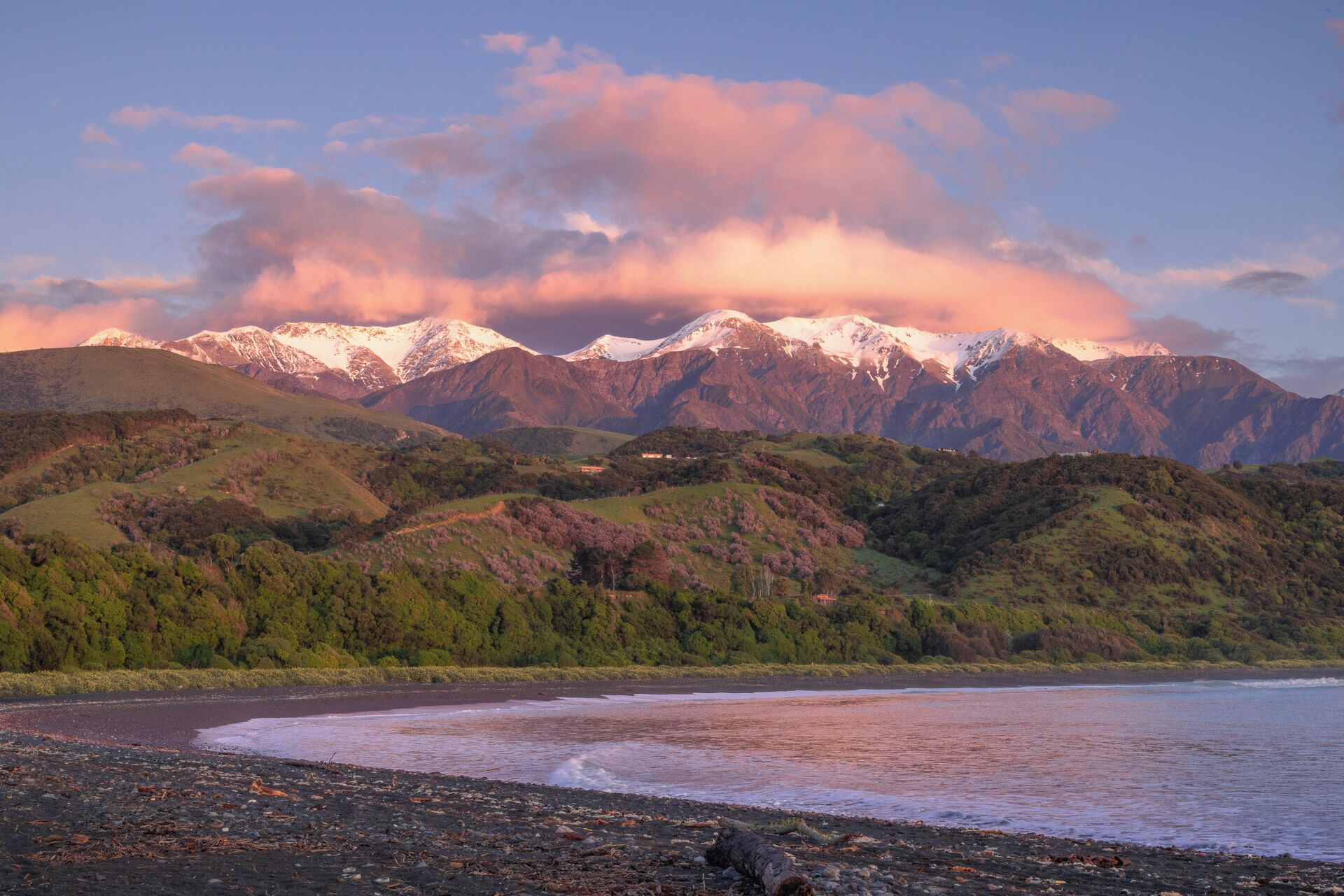 A coastal landscape with mountains in the background, a beach with pebbles, and a clear sky.