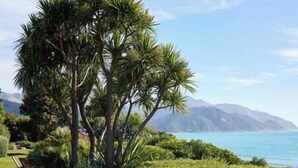 A tropical landscape with a palm tree, a chair, and a view of the ocean and mountains. - The Oceanview Suite (Hapuku)