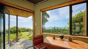 A room with a large window offering a view of a garden and the sea. There is a wooden table with a chair and a small table lamp. - The Oceanview Suite (Hapuku)