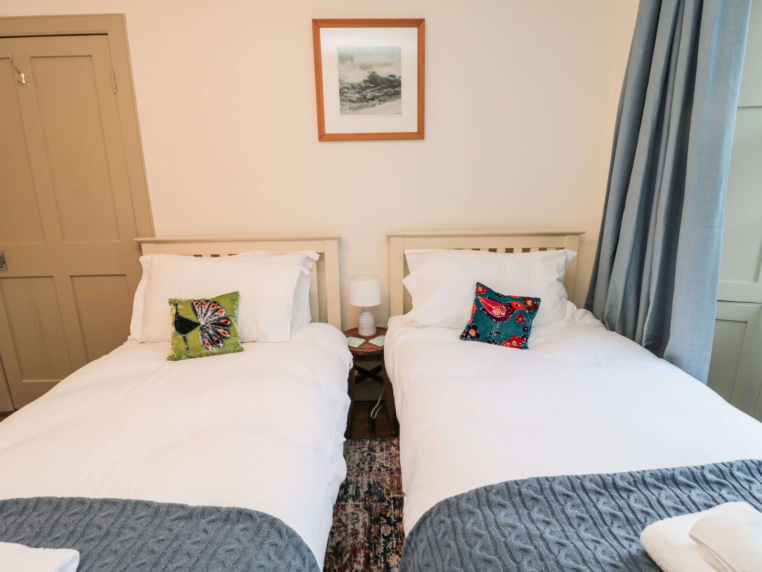 A hotel room with two single beds, each with a pillow and a blanket. There is a framed picture on the wall above the beds, a nightstand with a lamp, and a door to the left.