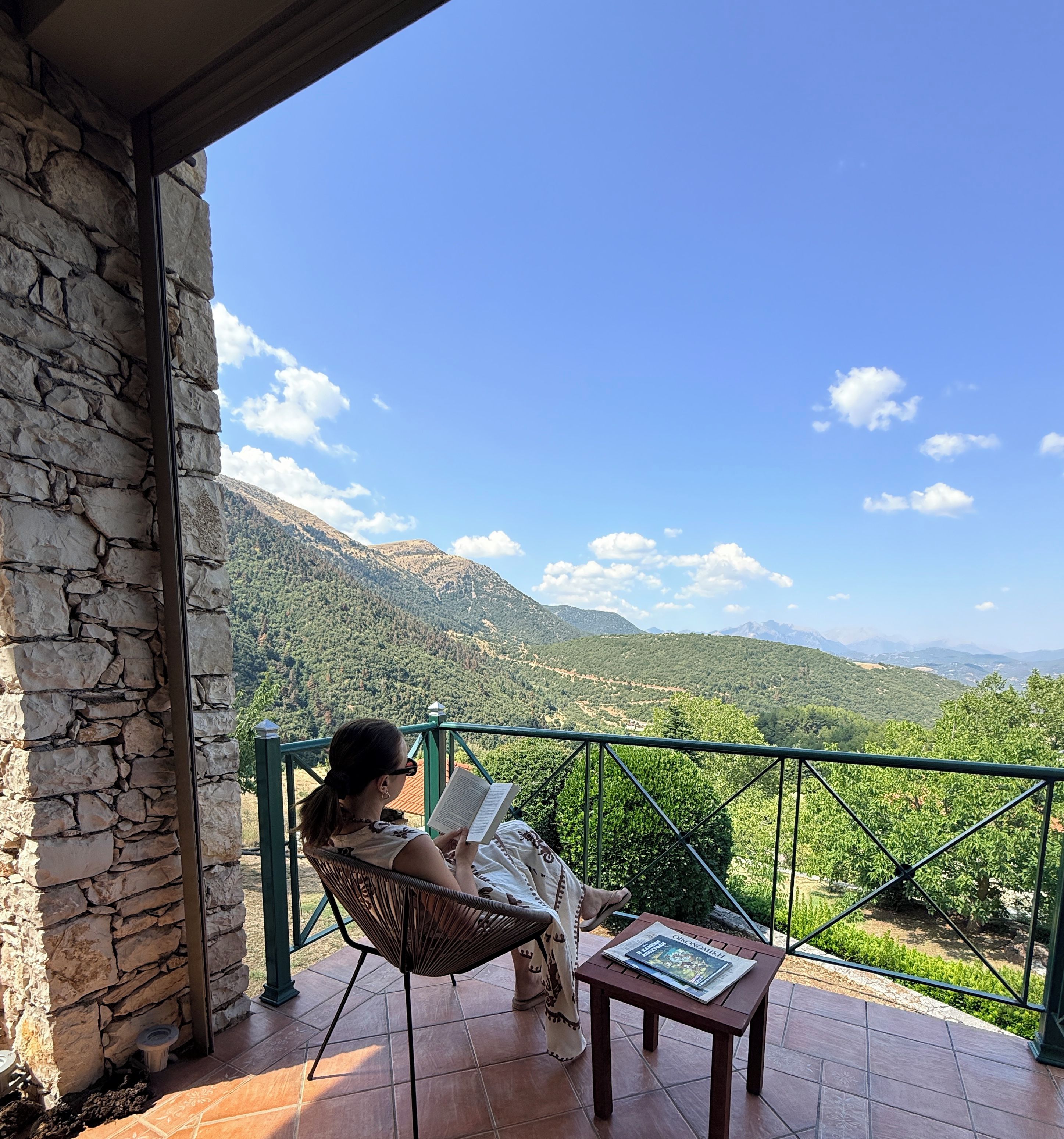 A person reading on a balcony with a view of mountains and greenery.