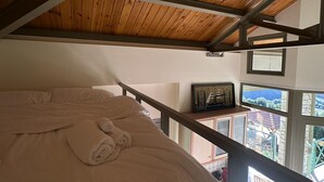 Ett sovrum med ett träpanelat tak, en säng med ett beige täcke och utsikt över ett berg genom ett fönster.