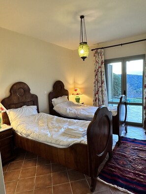 Une chambre avec deux lits en bois, une table de chevet avec une lampe, une fenêtre avec des rideaux et une vue sur l’extérieur.