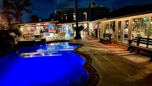 A swimming pool with a blue-lit edge, surrounded by a patio area with benches and a building with glass doors and windows. - Lake Charles #1 Rental located close to everything. Casino's/mall/restaurants.  (Lake Charles)