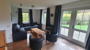 A modern living room with a sectional sofa, a wooden coffee table, and black armchairs. There are large windows with curtains, a clock on the wall, and a small wooden cabinet.